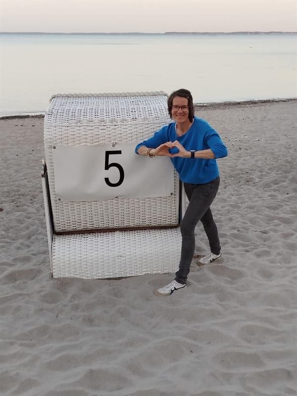 Andrea Heumann formt am Strand neben einem Strandkorb ein Herz mit den Händen