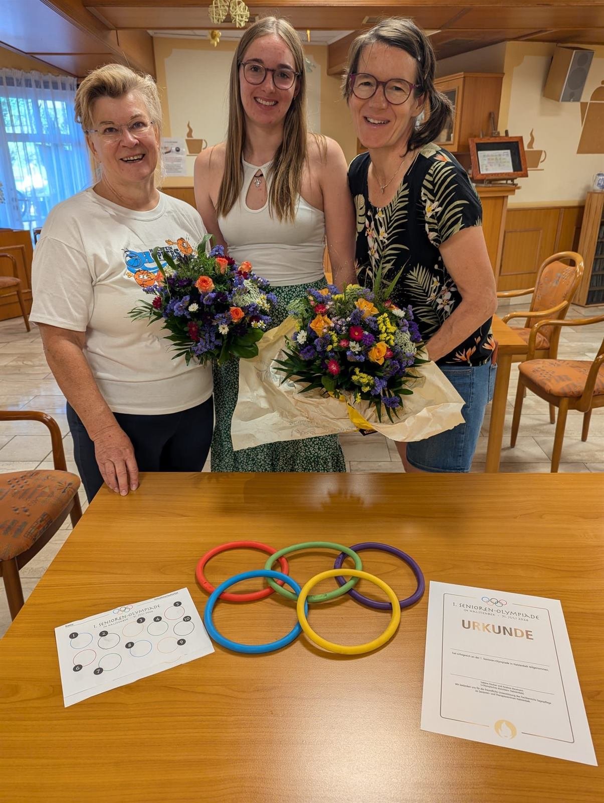 Drei Frauen mit Blumensträußen, davor bunte Ringe und eine Urkunde