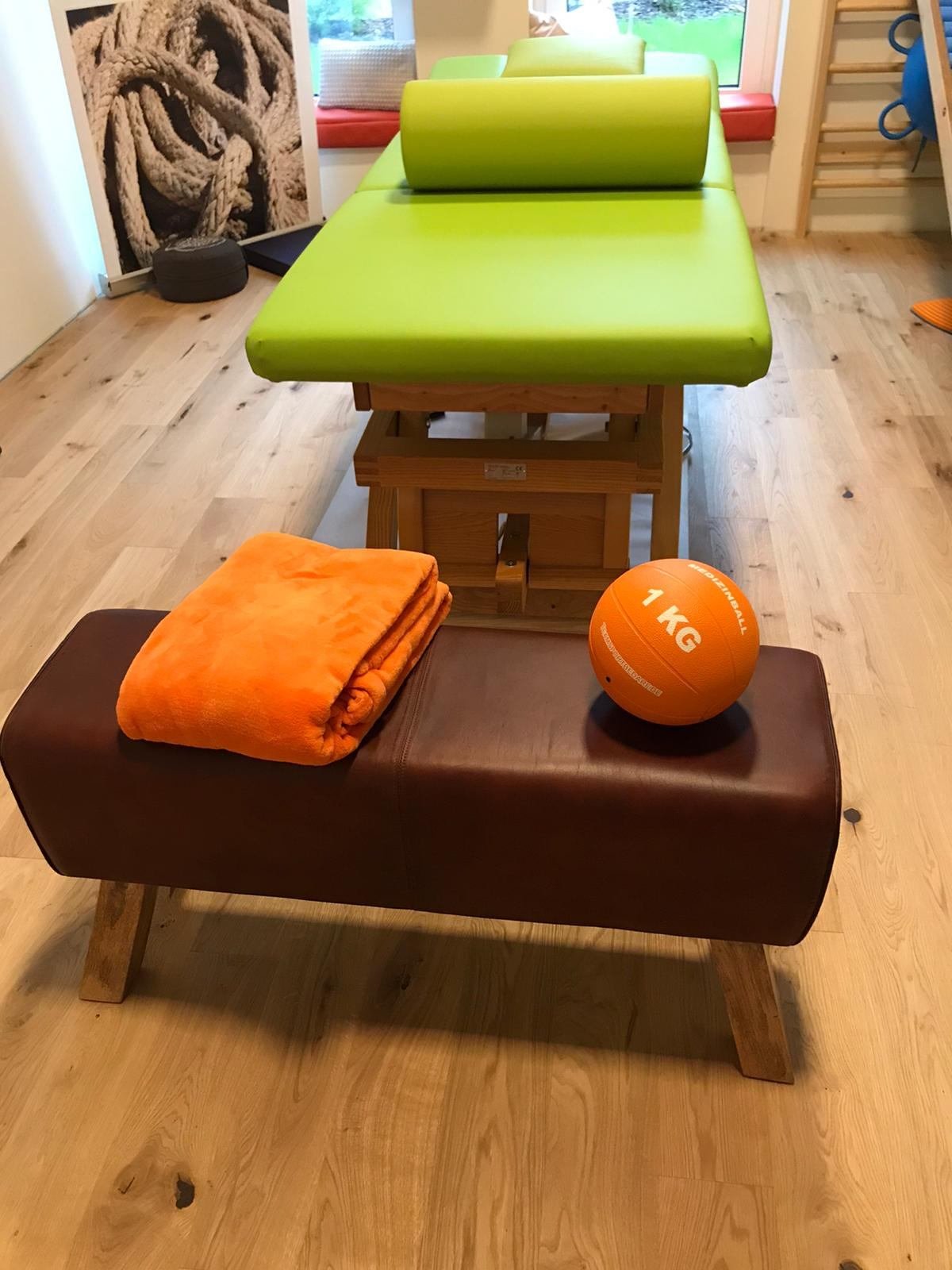 Grüne Behandlungsliege, davor ein Lederbock mit oranger Decke und Medizinball