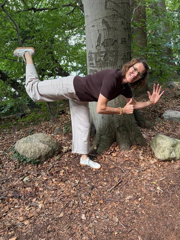 Andrea Heumann balanciert im Wald lachend auf einem Bein vor einem Baum