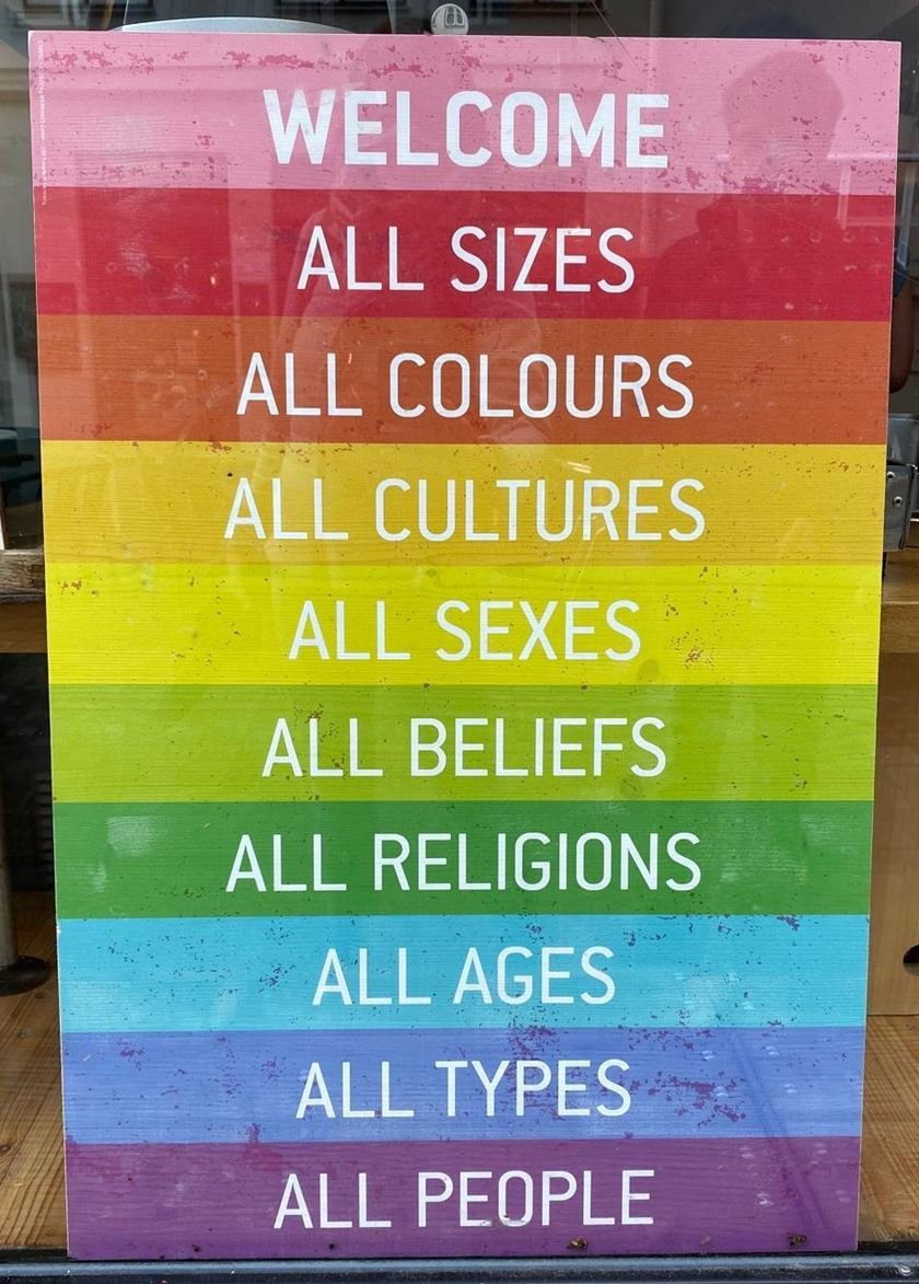 Schild in Regenbogenfarben: Welcome – all sizes, all colours, all cultures, all sexes, all beliefs, all religions, all ages, all types, all people